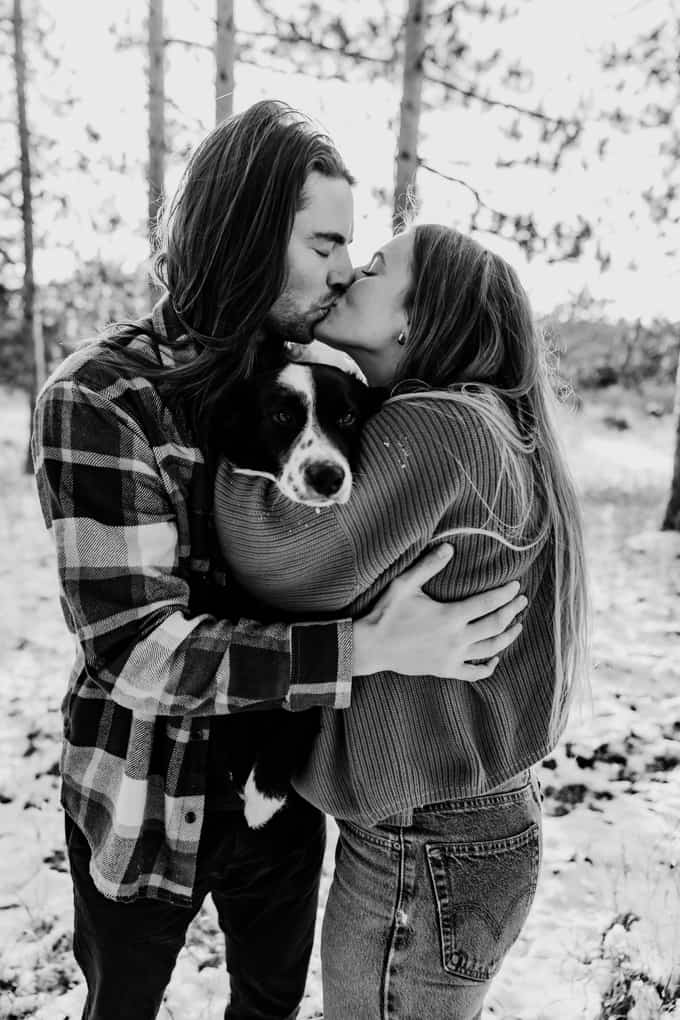 Couple hold their dog and kisses over his head during a winter golden hour