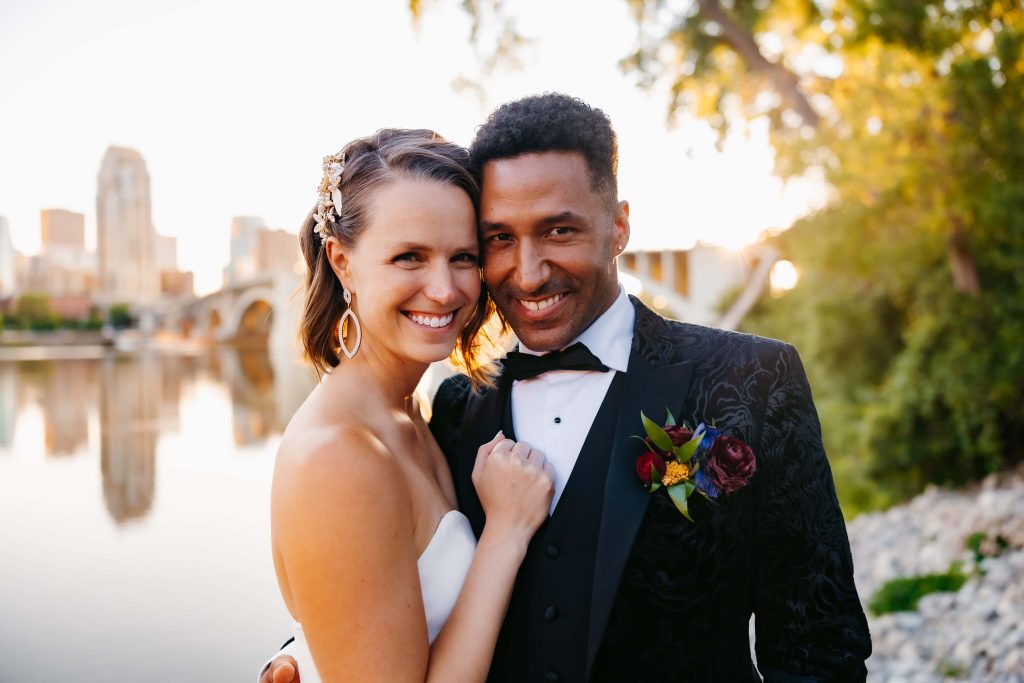 Caucasian bride stands with her African American husband in a textured black suit and they both smile at the photographer during their sunset wedding photos in Minnesota