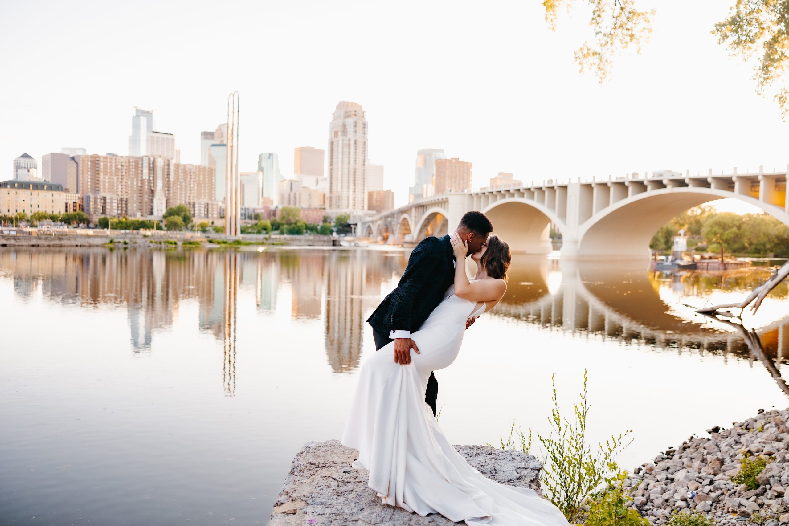 Groom in black suit dips his bride in a white dress for a kiss overlooking the Mississippi River