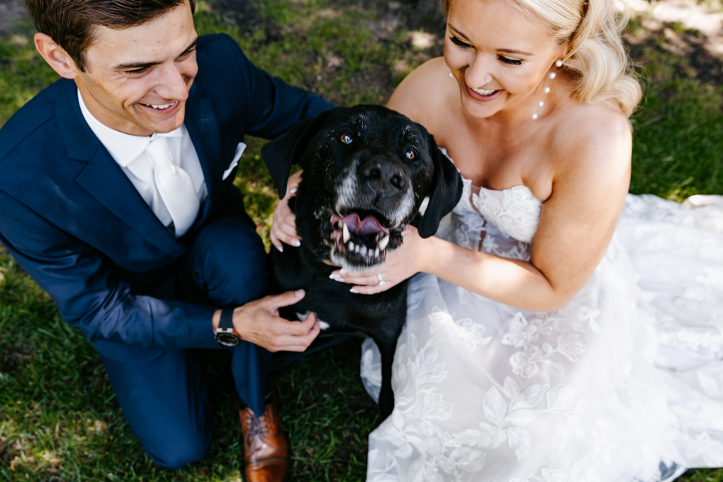 Couple squats down next to their black dog and smile at him at their Northern Oaks Events wedding