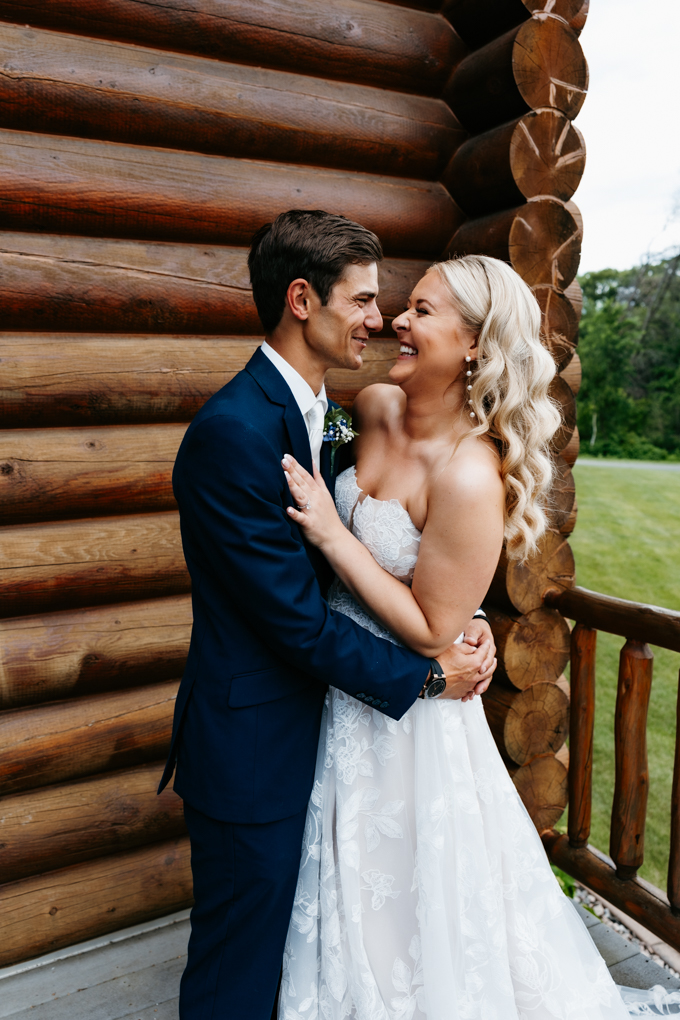 Bride and groom laugh together against a log cabin at their northern oaks event wedding 