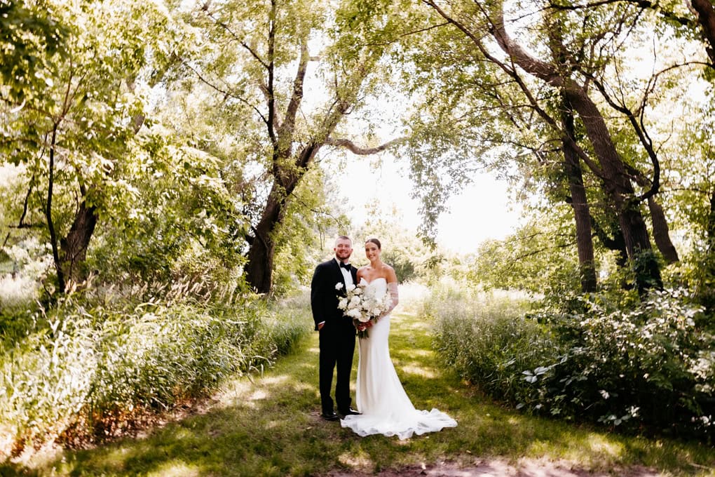 Bride in floor length white dress and groom in black suit stand together and smile at their Gathered Oaks Wedding