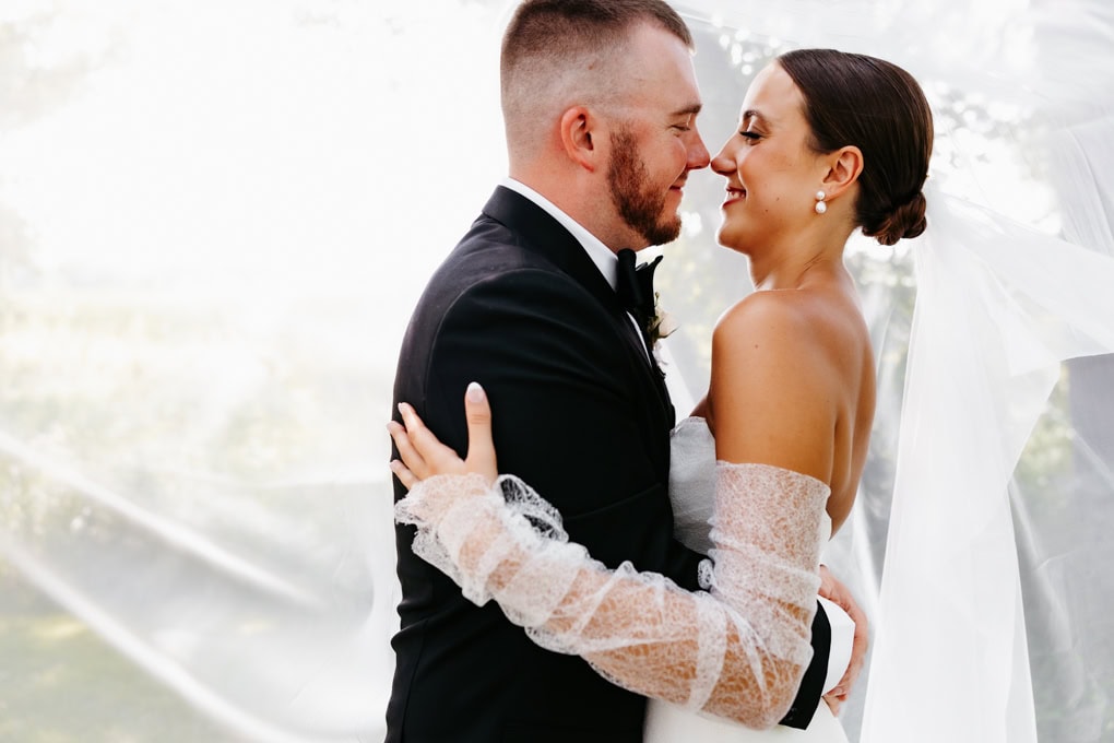 Bride and groom stand nose to nose under their veil at their gathered oaks wedding