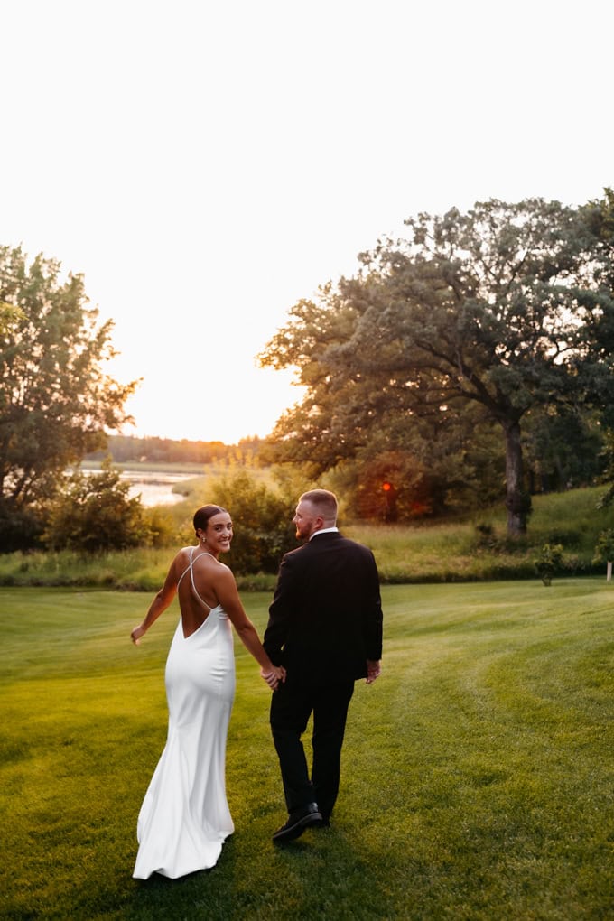 Bride in low back white dress holds hands with her groom in black tux as they walk down green lawn toward the lake at sunset on their Gathered Oaks wedding