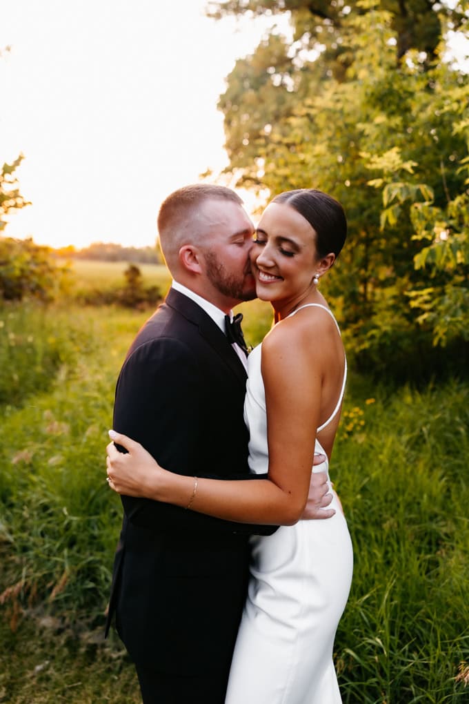 Bride in a white satin dress is held by her husband in black tux as he kisses her ear and she smiles at the Gathered Oaks Wedding