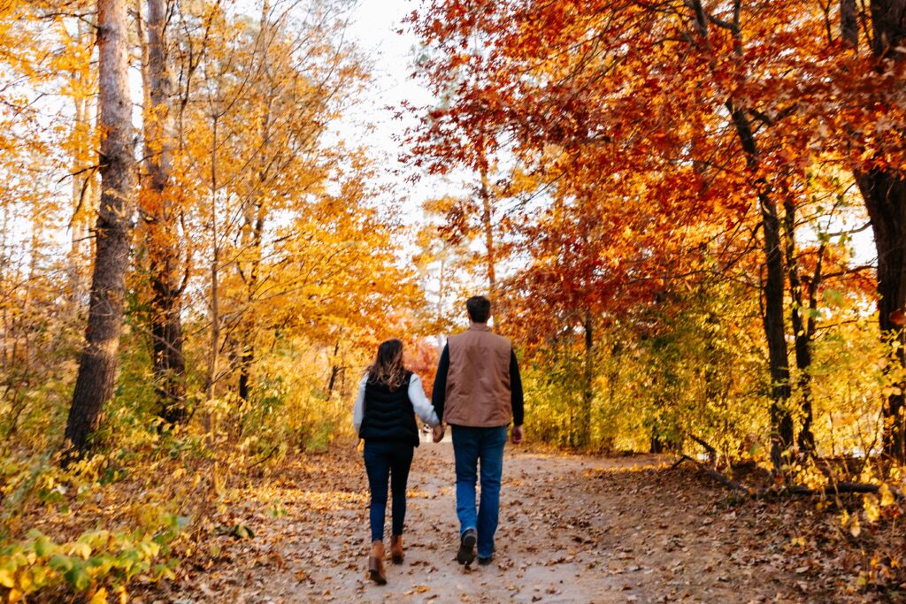 a man and woman holding hands walking on a path with trees in fall colors during their minneapolis engagement photography session