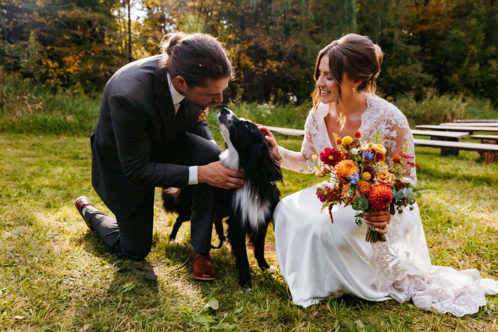 Groom in a green suit and bride in a white dress with bright orange, yellow and red flowers kneel down to pet their black and white dog during their 10-hour wedding timeline