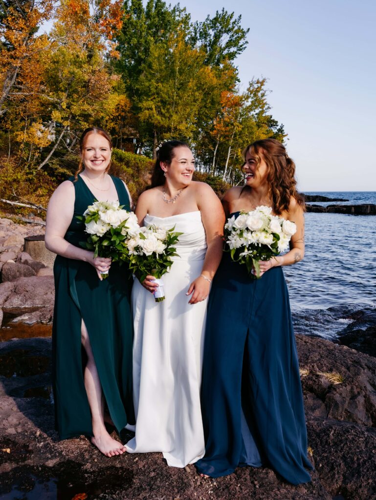 Bride in white dress stands between two white women with blue dresses on, all holding flowers and laughing on a rocky area in front of Lake Superior at the brides North shore wedding at Grand Superior Lodge