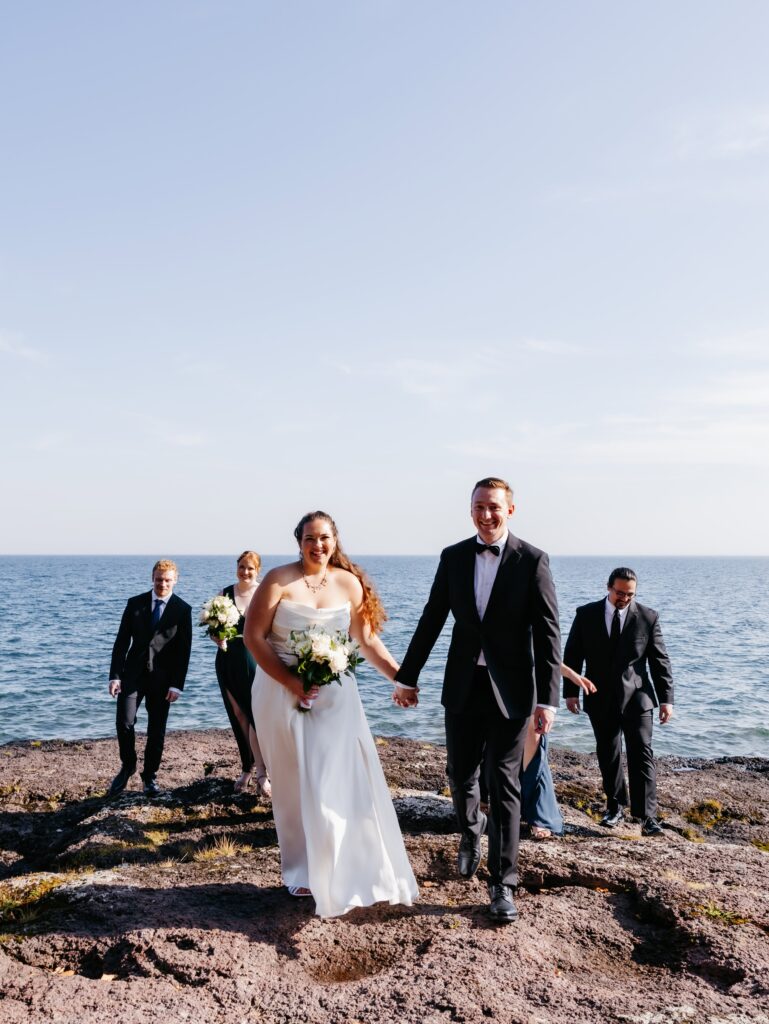 Bride and groom walk together smiling, bride holding flowers in her hand while a few men in tuxes and women in teal dresses walk behind them at SUperior SHores in Two Harbors MN