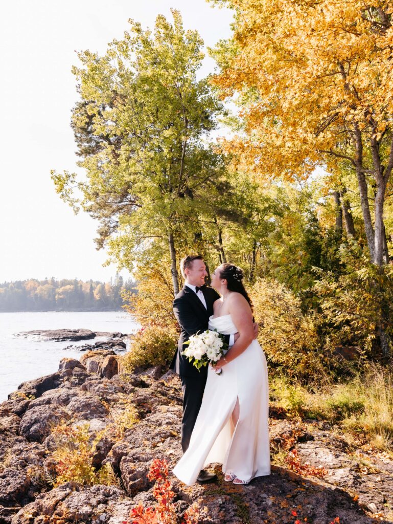 Bride in white dress with dark brown hair and groom in black tux hug and smile at each other on a rocky outcropping in front of tress that are green and orange in the fall at their SUperior shores wedding