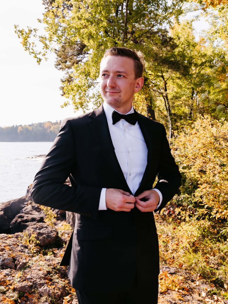 Groom in a black tux with black bowtie and white shirt smiles out to the left in front of lake superior