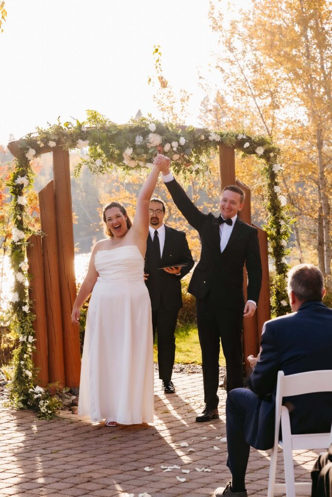 Bride and groom hold hands in front of a wooden arch and cheer after they got married at Grand Superior Lodge