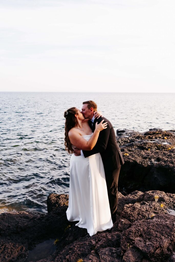Bride and groom embrace and kiss on a rock in front of Lake Superior at their Grand Superior Lodge wedding