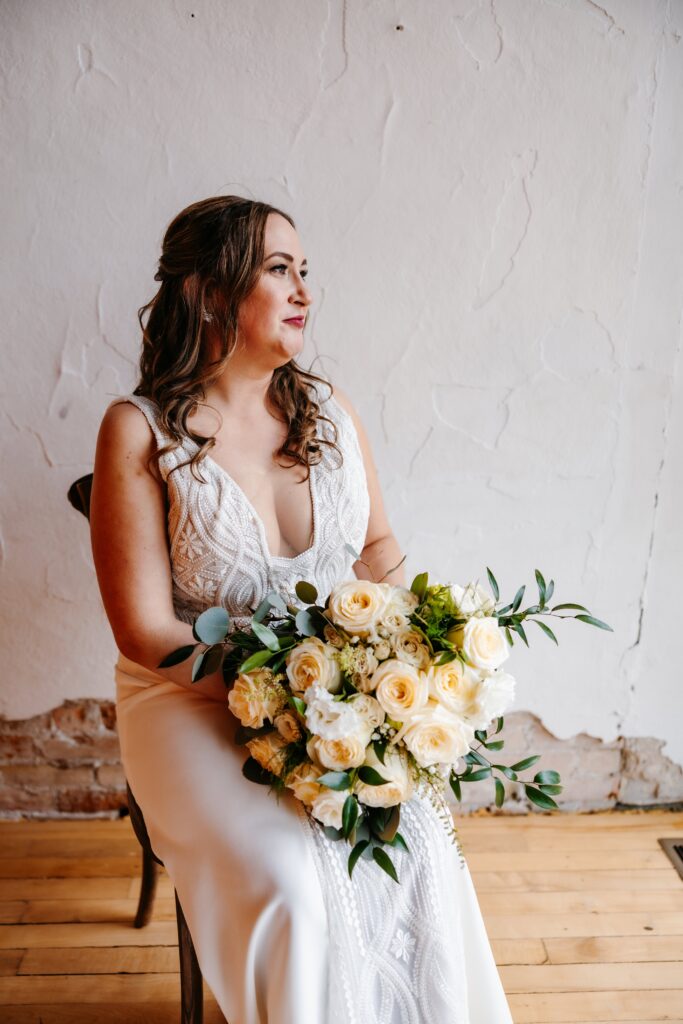 Bride in a v neck white dress holds her white and yellow bouquet and stares out a window at her jerome event center wedding