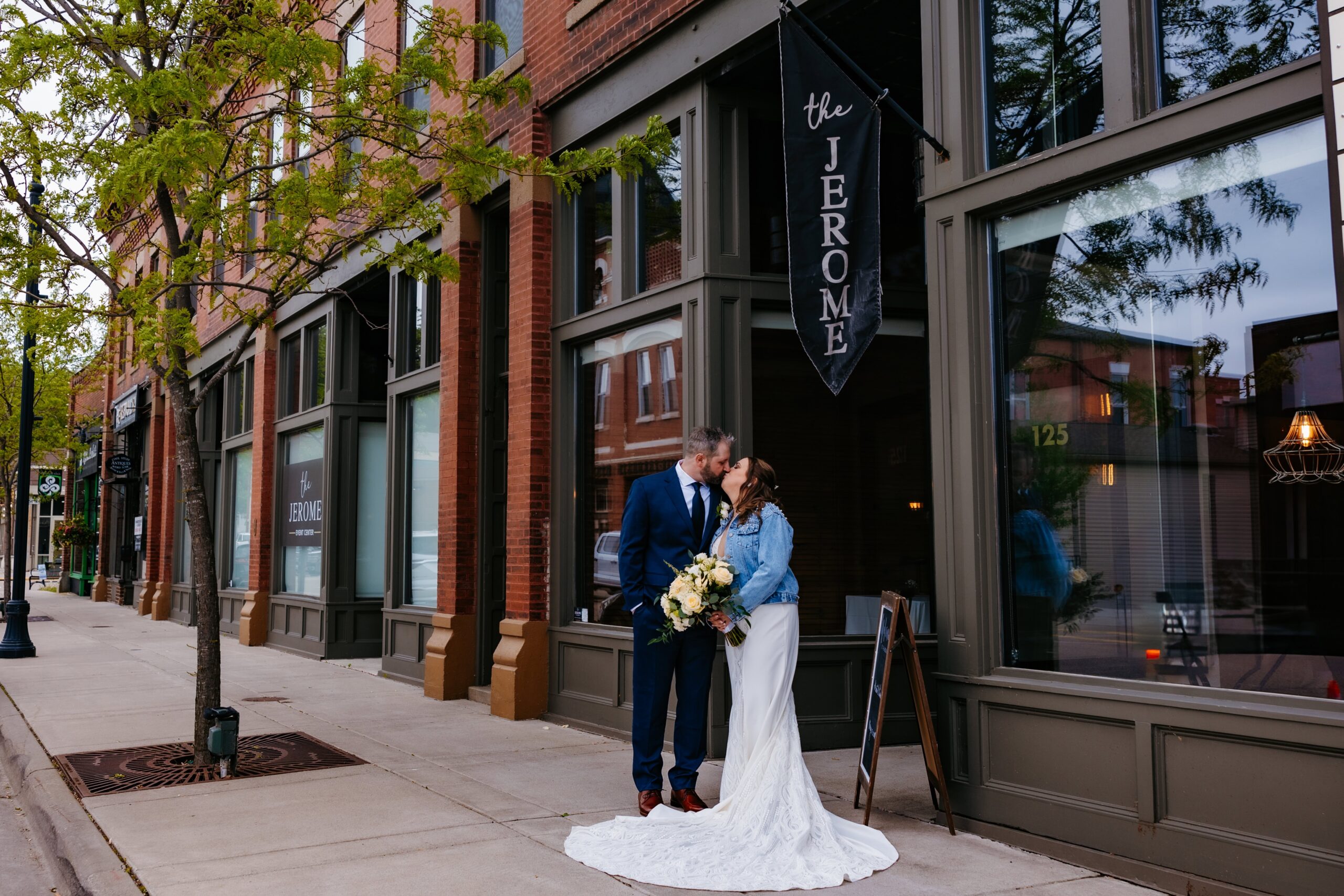 A bride in a white dress and jean jacket stands with her groom in a suit on the outside of the brick Jerome Event Center wedding