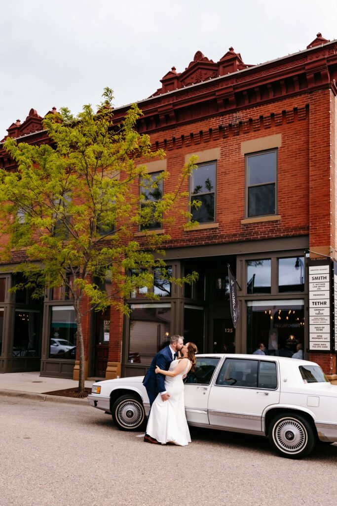 Bride and groom kiss in front of an old white car in front of the venue for their Jerome Event Center wedding