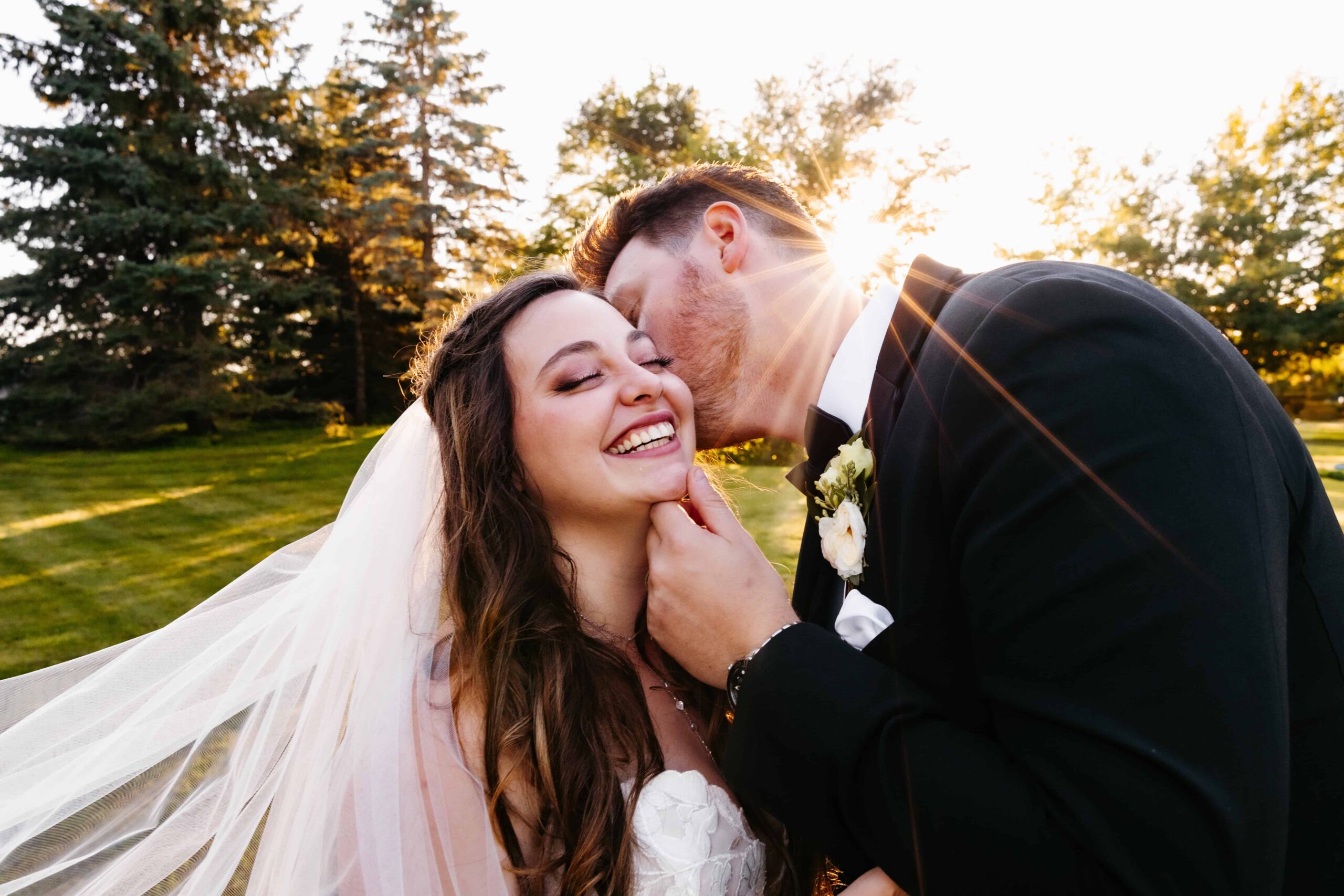 Groom in a black tux cups his wife with brown hair's face as she smiles at sunset at her gardens of castle rock wedding